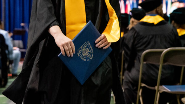Graduating student carries a blue diploma cover with The College of Saint Scholastica official shield.