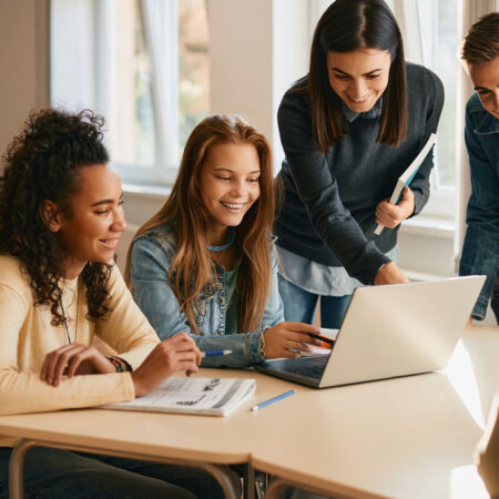 Teacher assisting a group of students with an assignment on a laptop in a classroom.