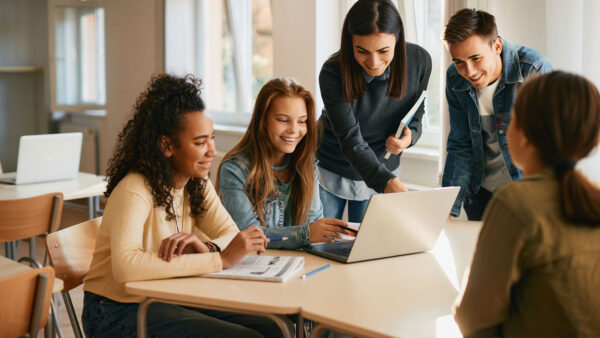 Teacher assisting a group of students with an assignment on a laptop in a classroom.