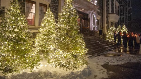 Students, faculty and staff gathering on the steps of Tower Hall in front of fully decorated holiday trees.